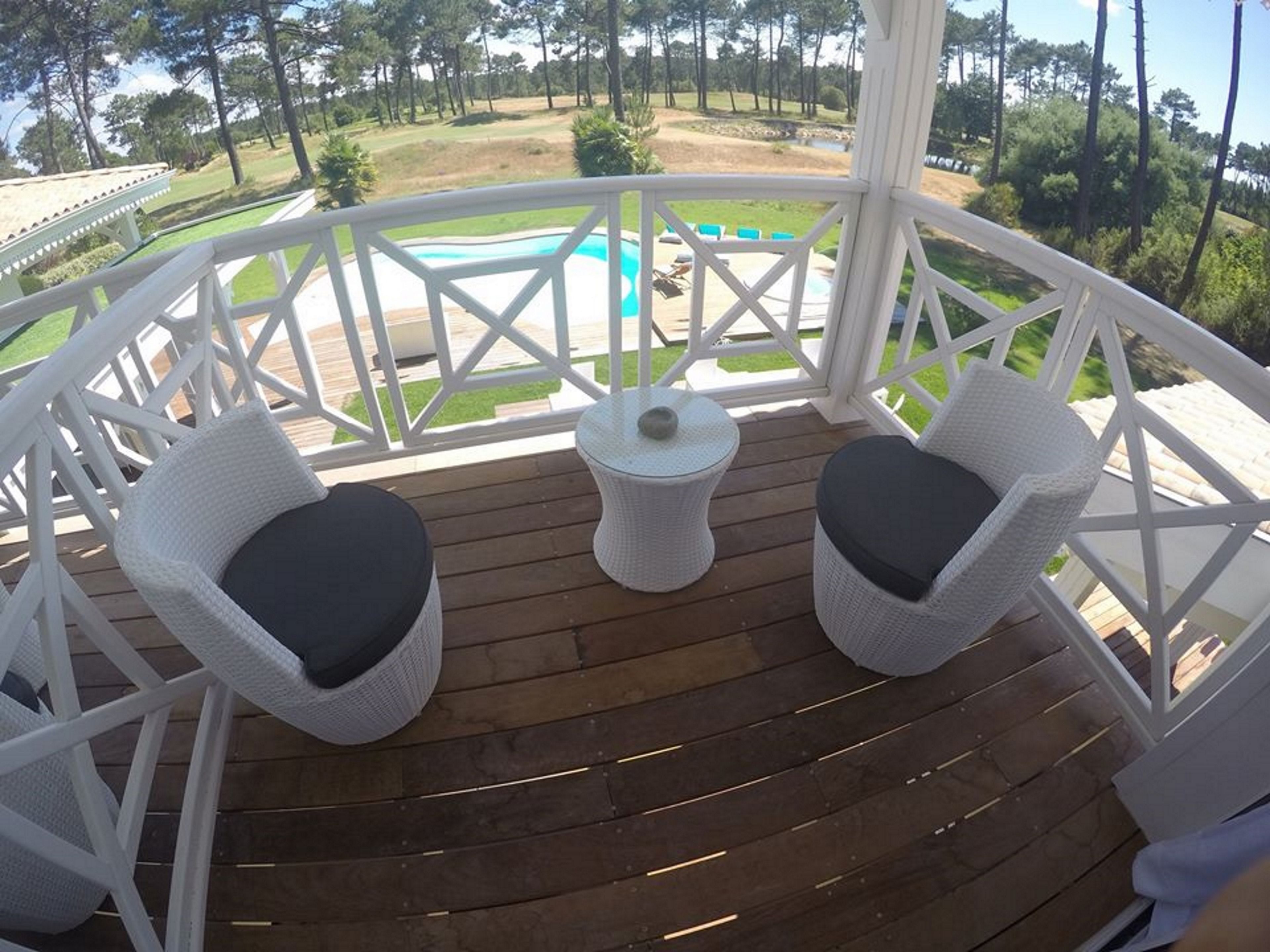 Ett takförsett utomhusområde med två vita flätade fåtöljer med mörka kuddar, ett litet runt bord med en vit bordsskiva och en enda mörk kudde, samt utsikt över en golfbana och en swimmingpool i bakgrunden.
