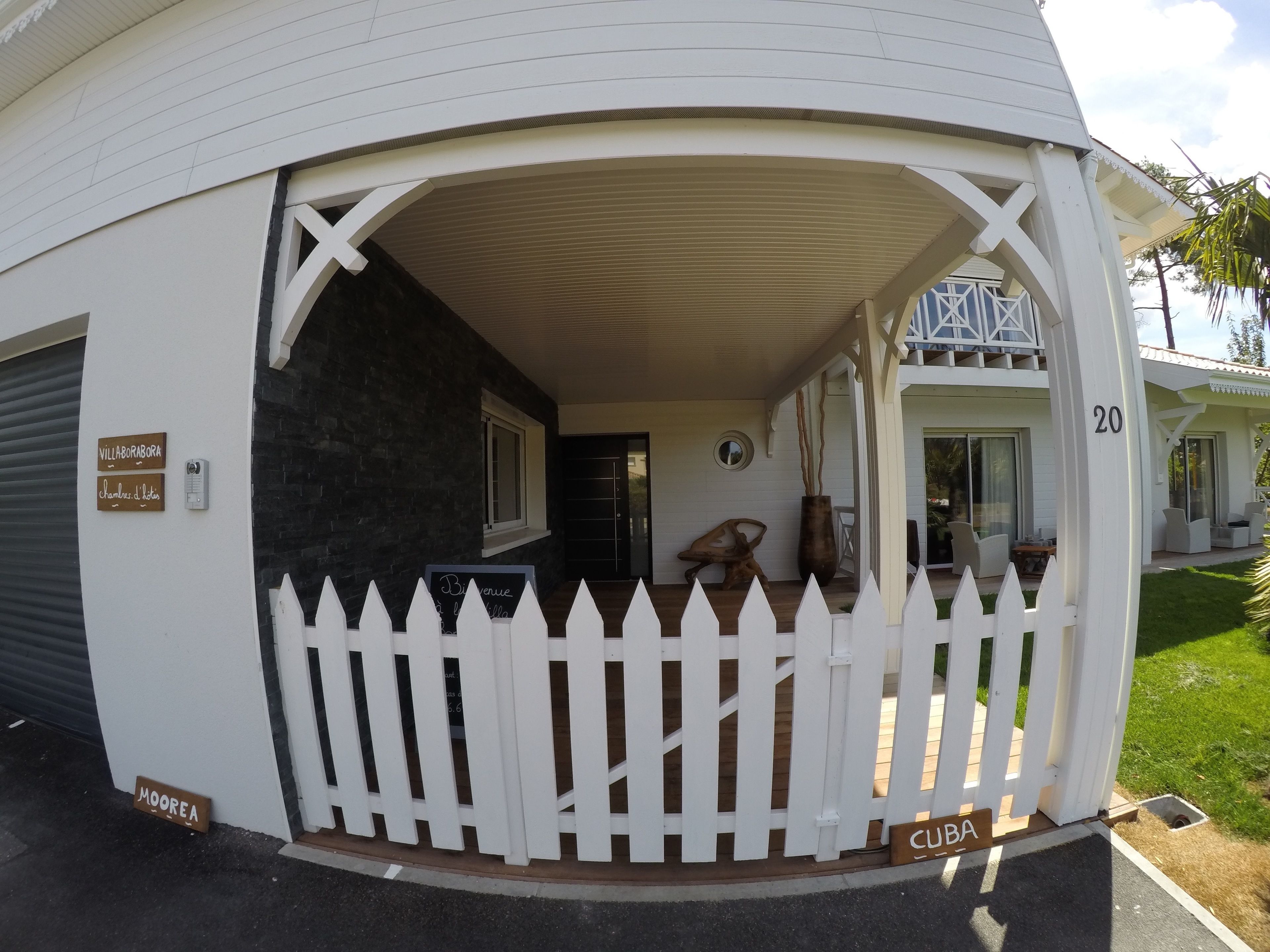 A white building with a covered porch, a white picket fence, and a number '20' sign.