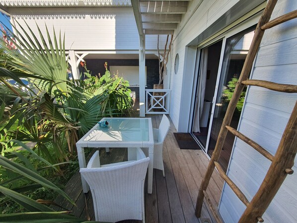 A covered patio area with a glass table and wicker chairs, surrounded by lush greenery and a wooden deck.