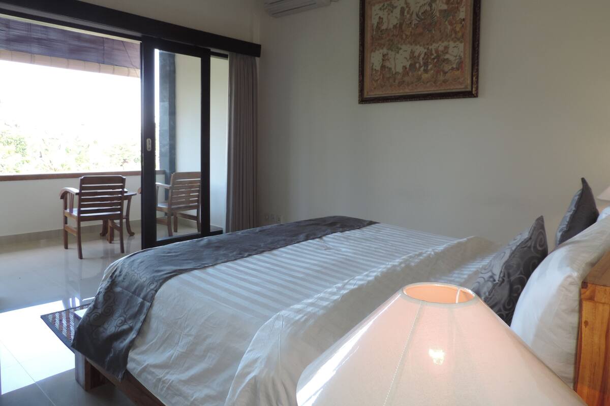A bedroom with a bed, a chair, a table, a window, and a painting on the wall.