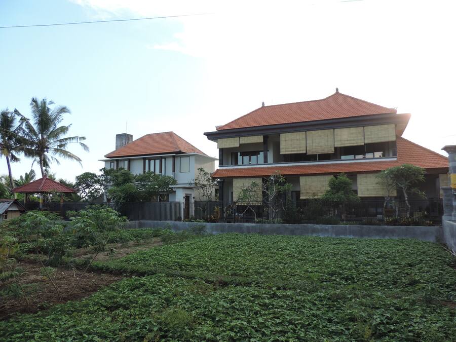 Une maison à deux étages, avec un toit recouvert de tuiles rouges, entourée de verdure et dotée d’un jardin clos.
