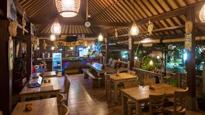Un restaurante interior con mesas y sillas de madera, luces colgantes y vistas a un jardín exterior.
