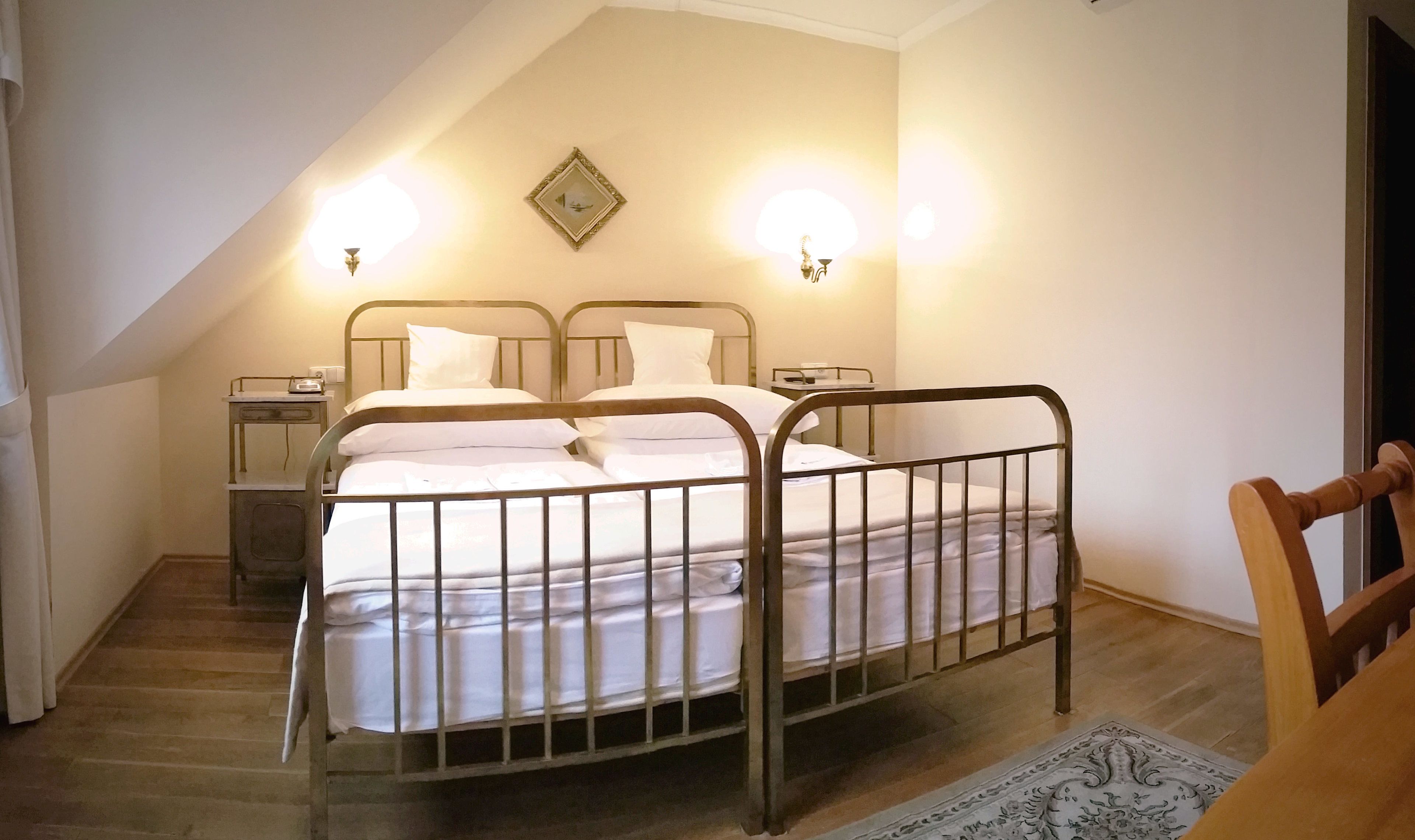 Quarto com estrutura de cama de metal, duas mesas de cabeceira, um criado-mudo e um aparelho de luz fixado na parede.
