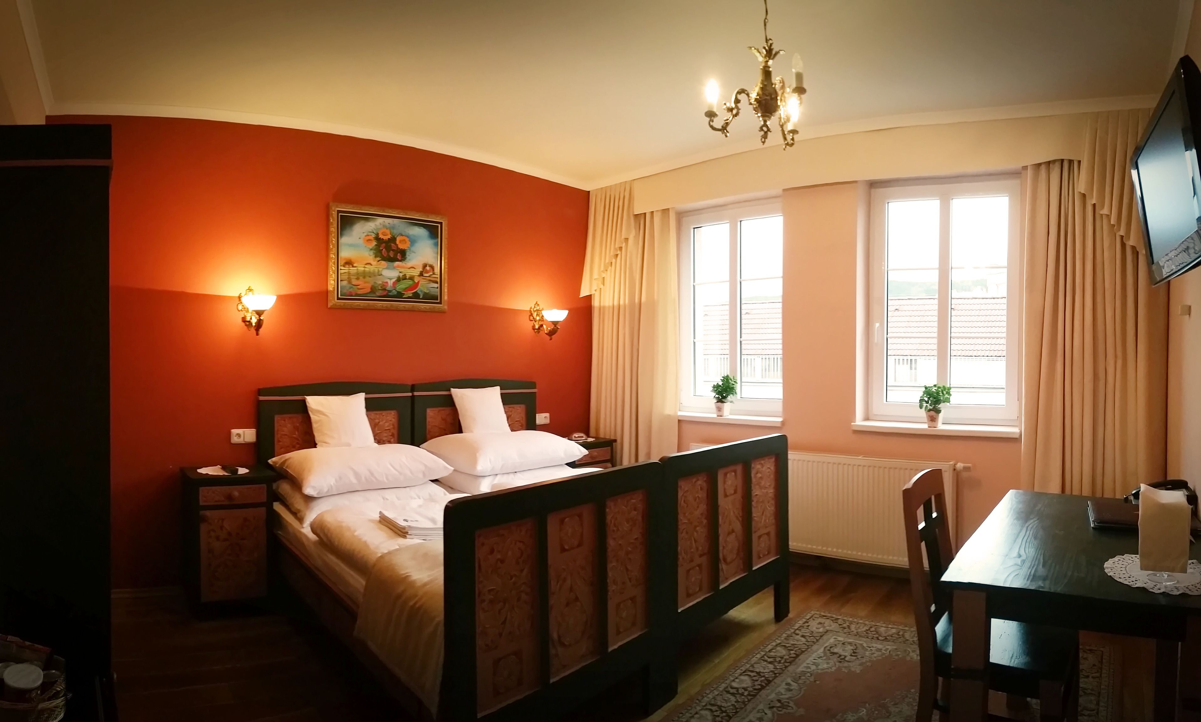 A bedroom with a double bed, two bedside tables, a desk with a chair, a chandelier, a painting on the wall, and a window with curtains.
