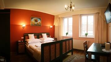 A bedroom with a double bed, two bedside tables, a desk with a chair, a chandelier, a painting on the wall, and a window with curtains.