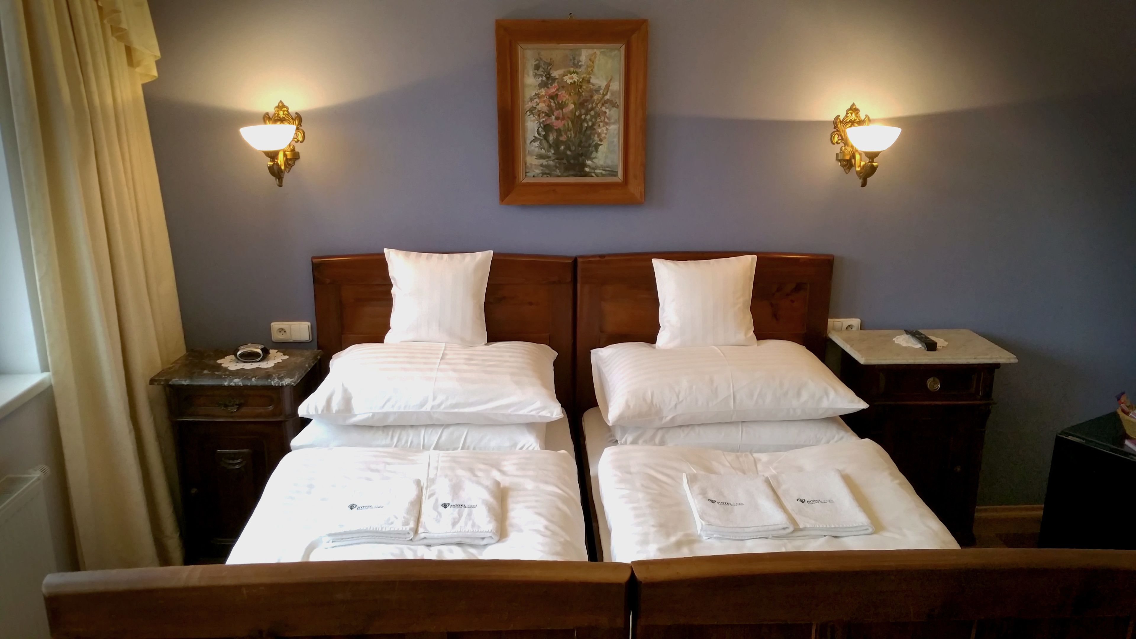 A hotel room with two beds, a nightstand with a lamp, a painting on the wall, and a window with curtains.