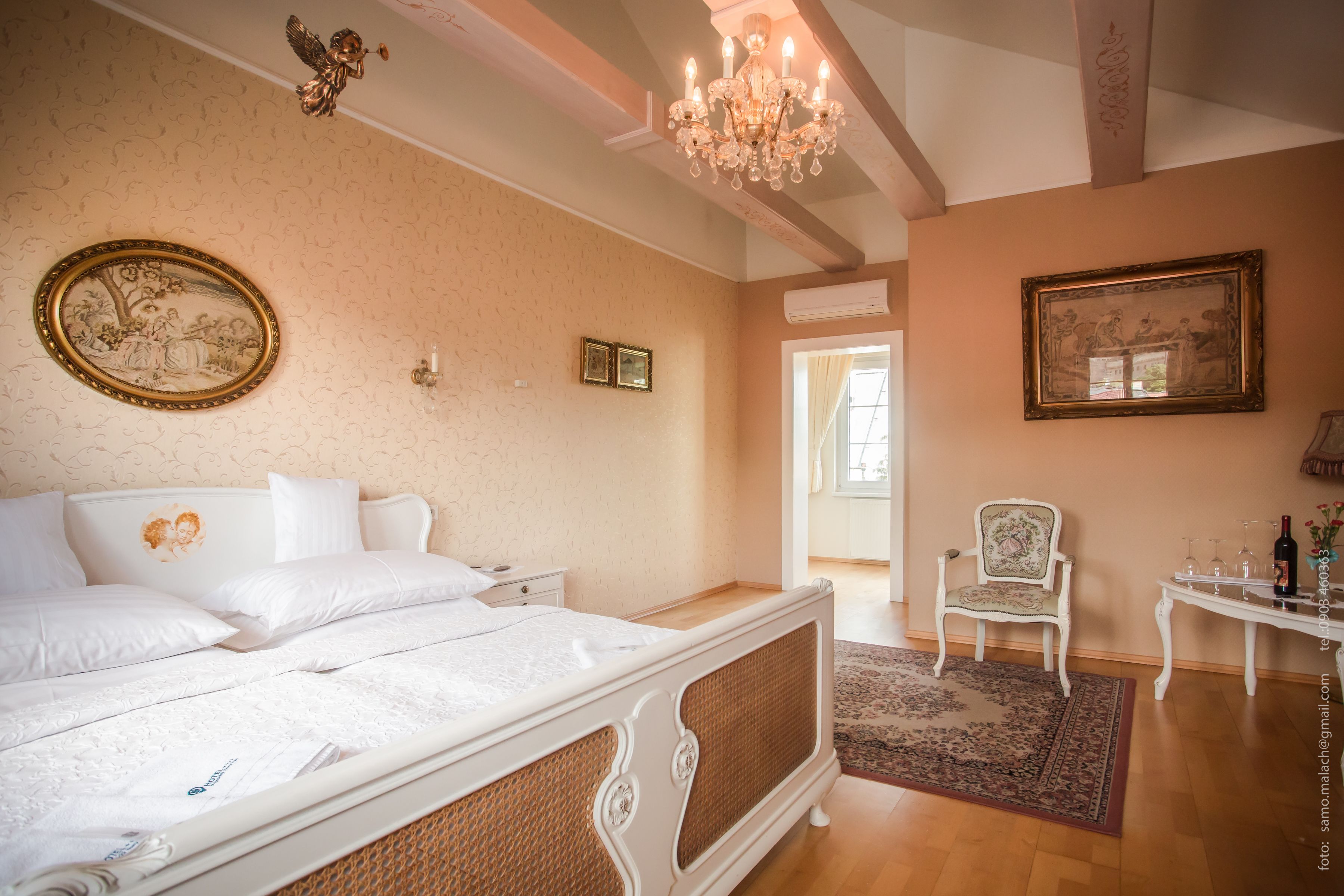 A bedroom with a large bed, a chandelier, a chair, and a table with glasses and a bottle.