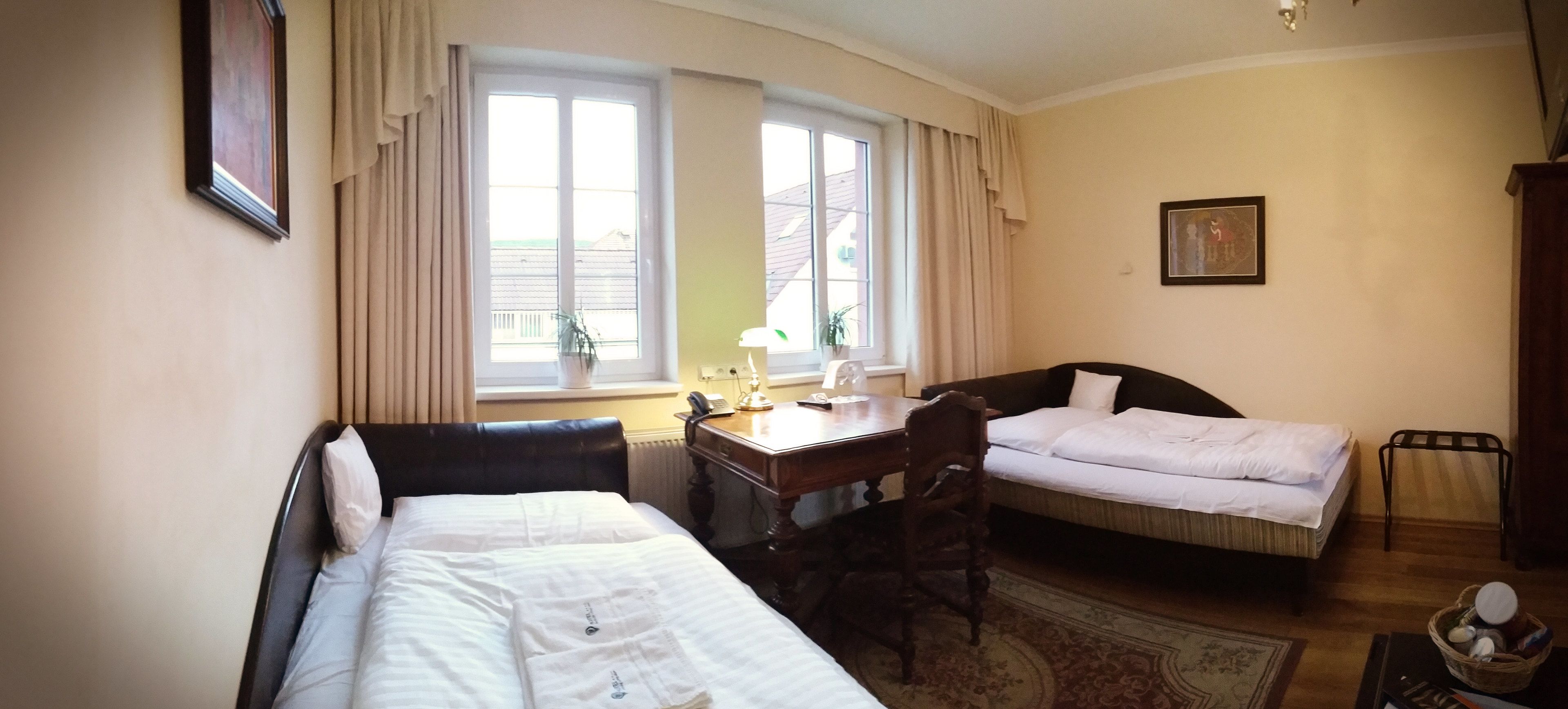 Une chambre d’hôtel avec deux lits, un bureau, une chaise, une fenêtre avec des rideaux, une plante et un tableau au mur.