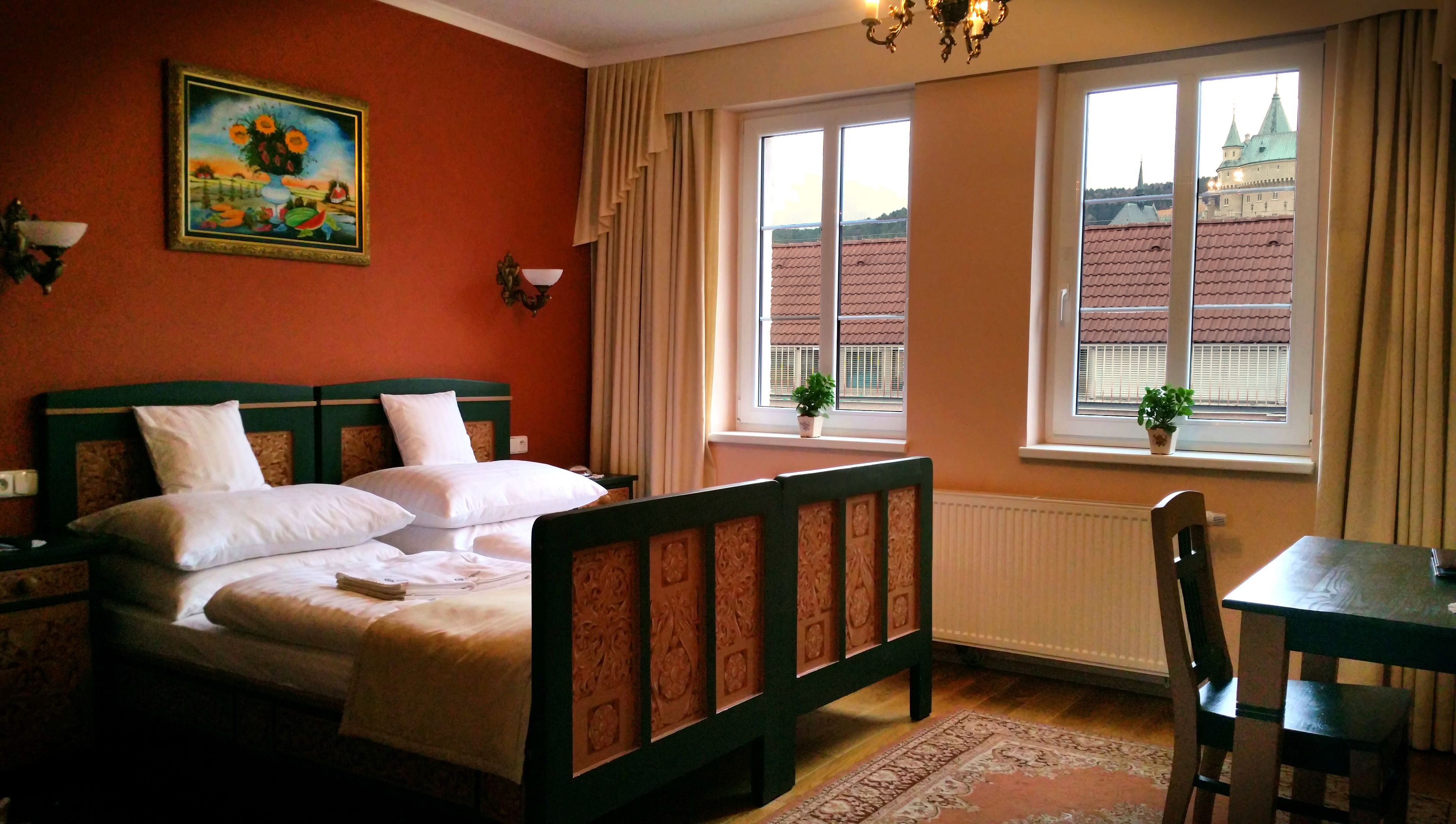 A room with a bed, a table, and a chair. There is a window with a view of buildings outside.