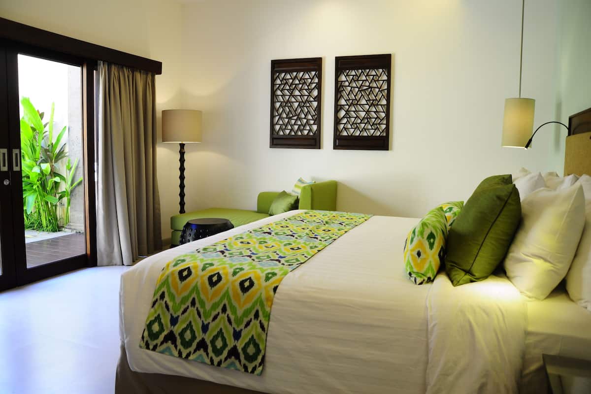 A hotel room with a bed, a green armchair, a small table, and a view of greenery outside.