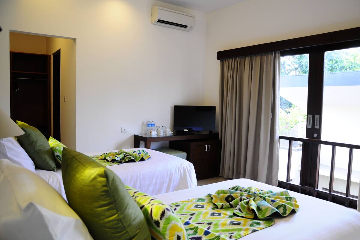 A hotel room with two beds, a TV on a wooden cabinet, an air conditioning unit, and a balcony with curtains.