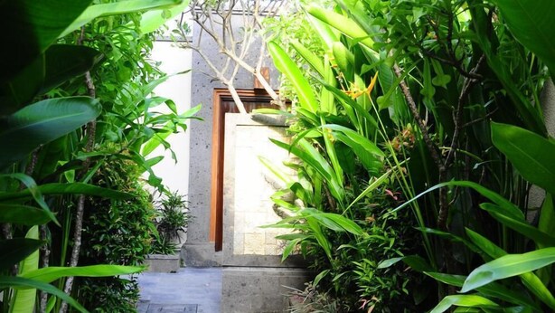 A narrow stone pathway flanked by lush green plants leading to a wooden door. - Puri Mesari Suites (Denpasar)