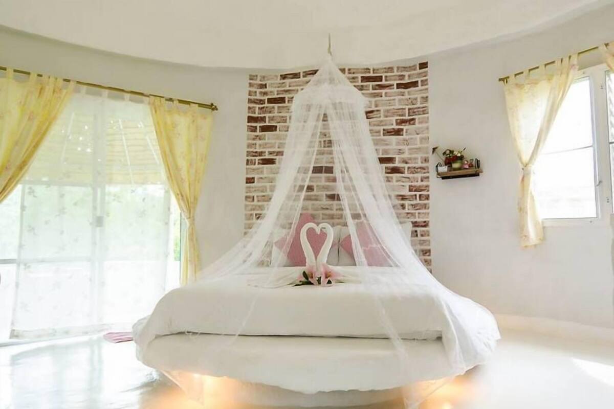 Een slaapkamer met een bed onder een witte hemelpartij, een muur met bakstenen motief en een raam met gele gordijnen.