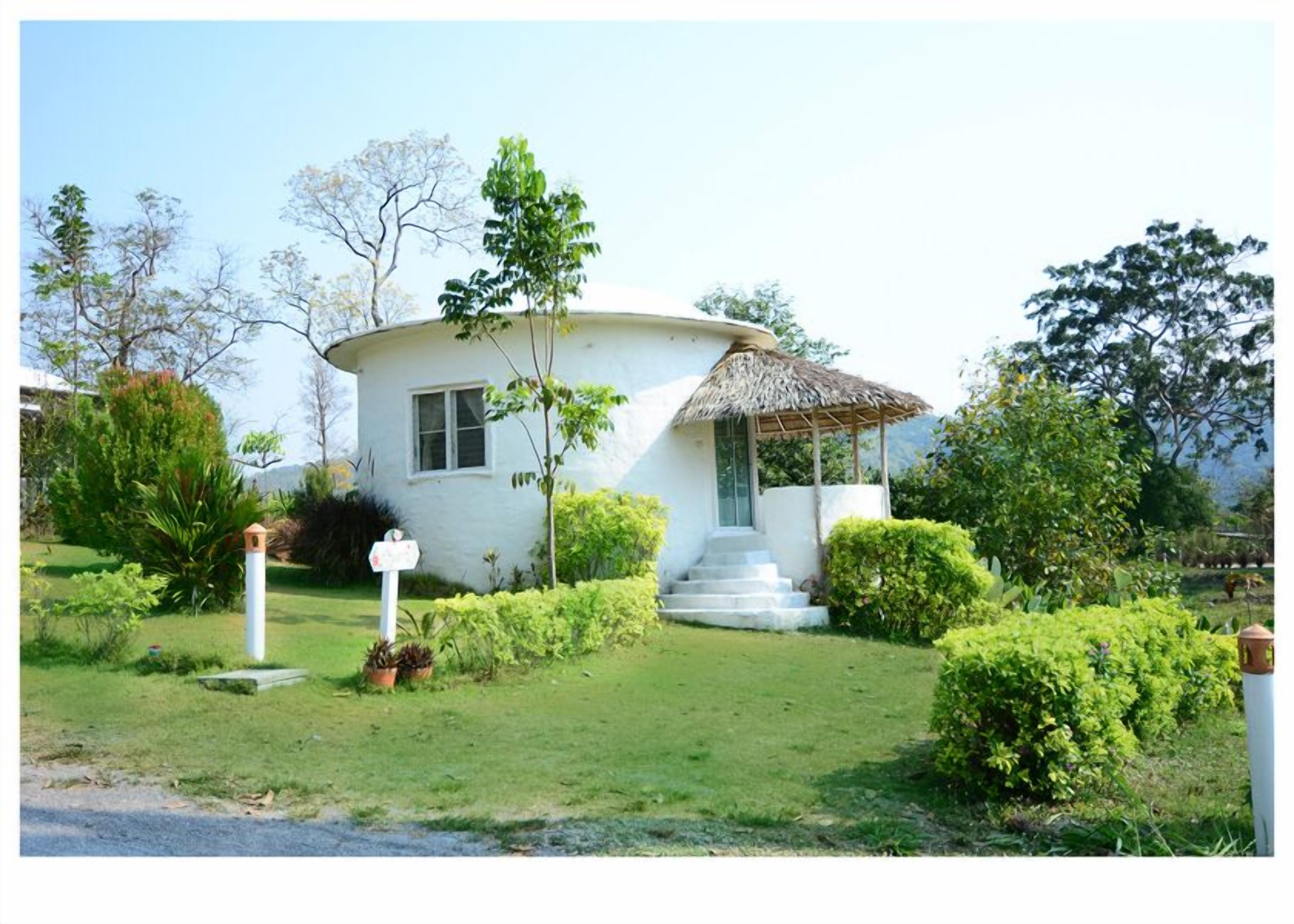 A white, single-story house with a thatched roof porch, surrounded by well-maintained greenery and a clear sky.