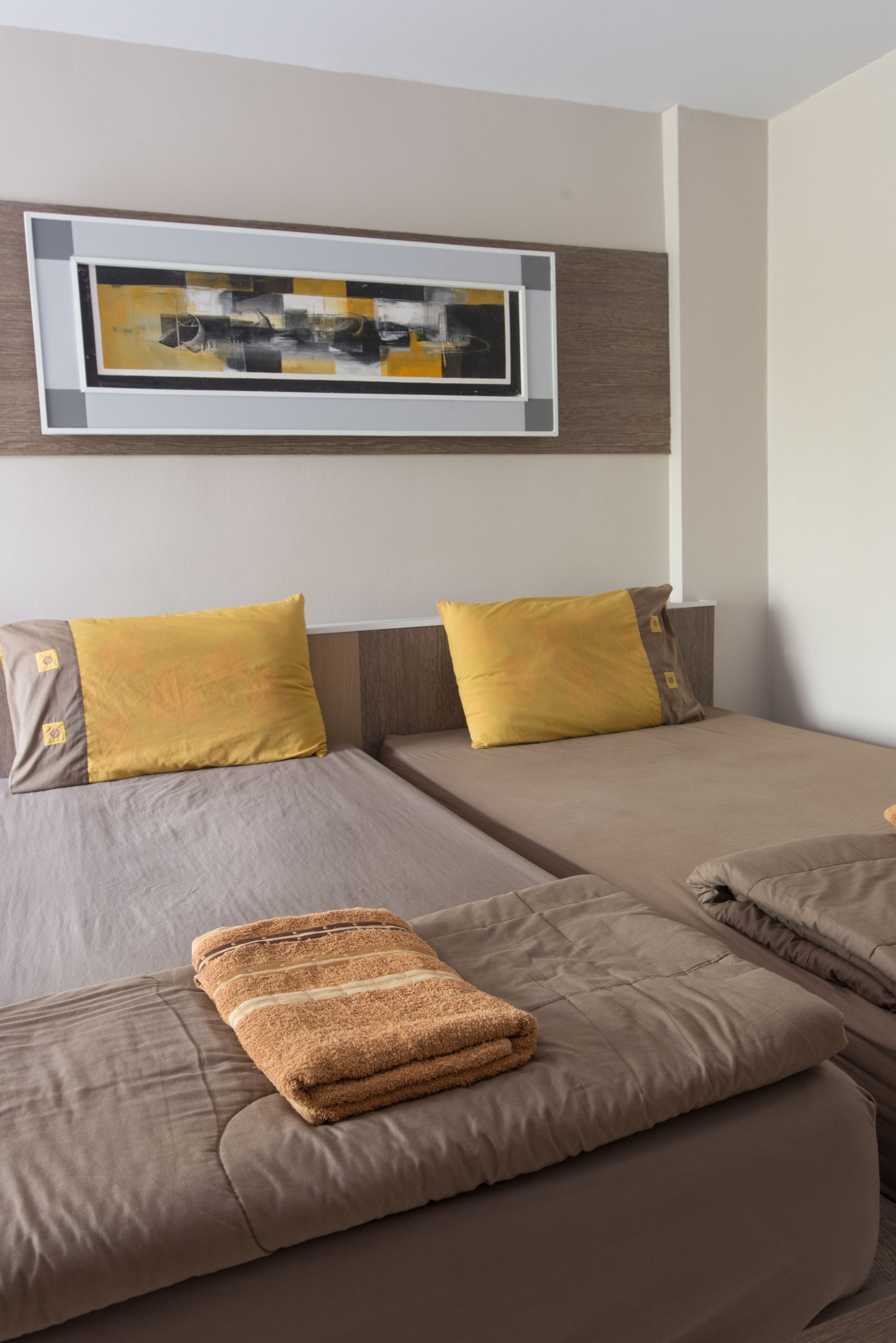 Habitación de hotel con dos camas, cada una con una colcha marrón y una almohada amarilla. Una toalla marrón doblada está sobre la cama inferior.