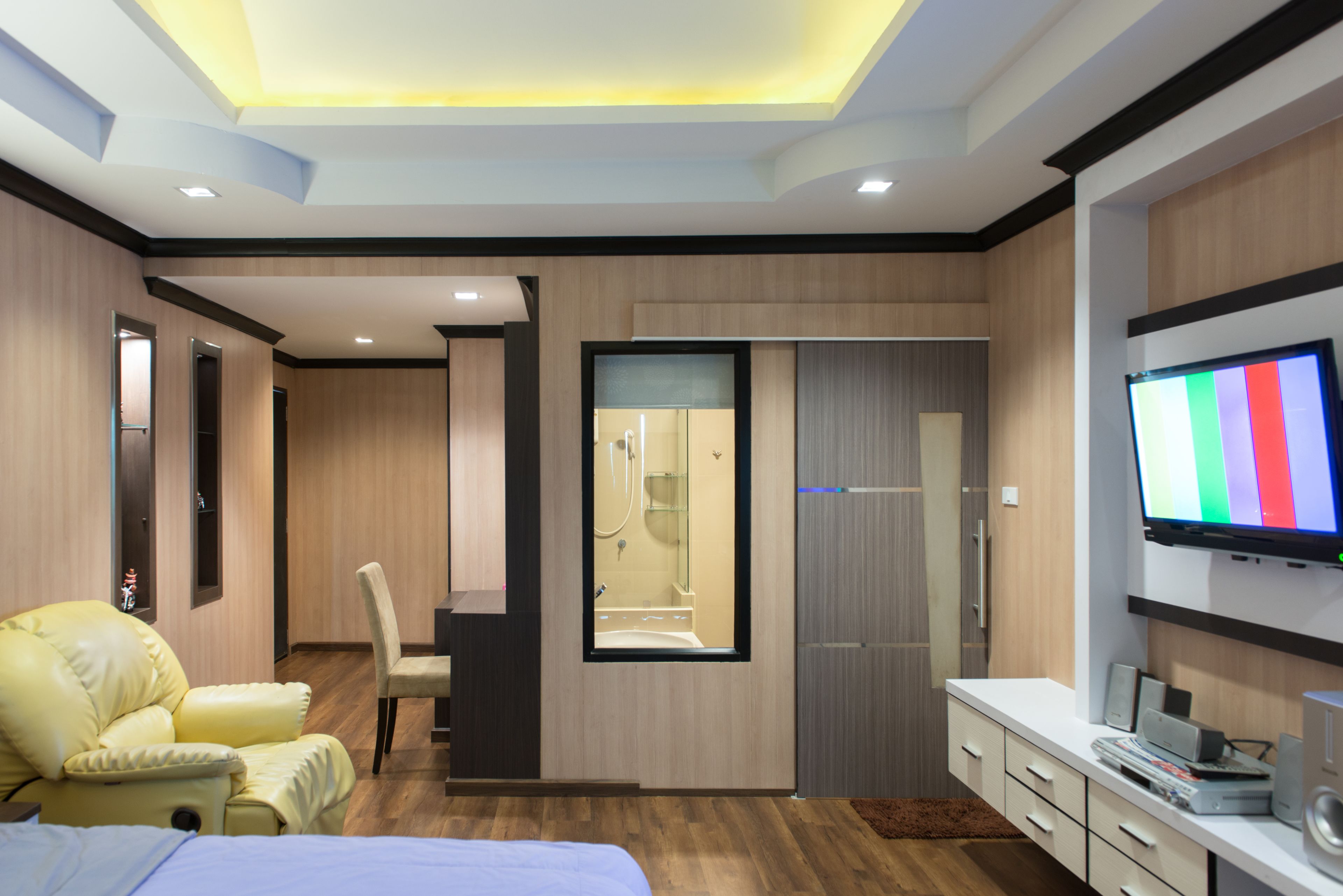 Una habitación de hotel moderna con cama, un sillón amarillo, televisión y baño con espejo y ducha.