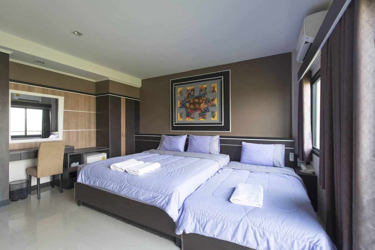 Habitación de hotel con una cama grande, un escritorio y una silla. Hay un cuadro en la pared y un equipo de aire acondicionado sobre la cama.