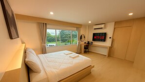 Un dormitorio moderno con una cama grande, un escritorio, un televisor y vistas al exterior con vegetación.