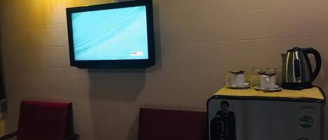 A room with a television mounted on the wall, a small table with a remote control, and a refrigerator with a kettle and cups on top.