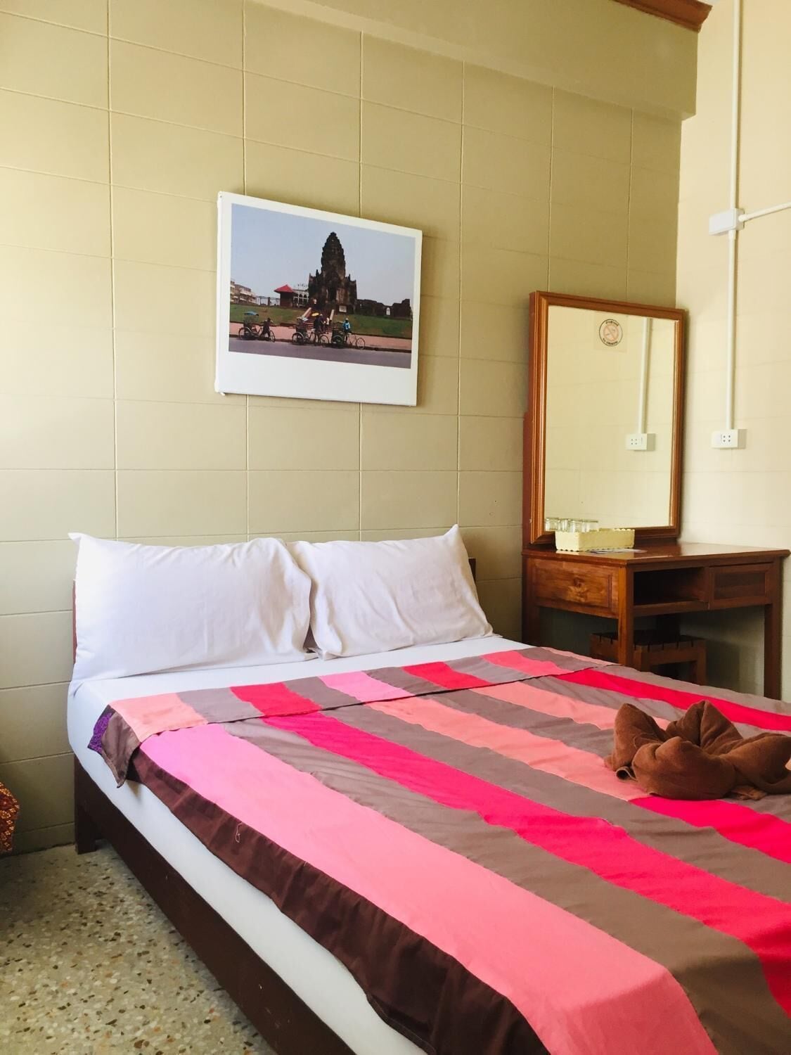 Una habitación de hotel con una cama, una mesita de noche de madera, un espejo y un cuadro colgado en la pared que representa un templo.