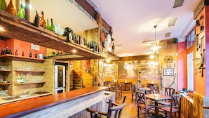 Uma área de bar rústica com prateleiras de madeira exibindo garrafas e copos, balcão de madeira e um ambiente acolhedor com cadeiras e mesas de madeira.