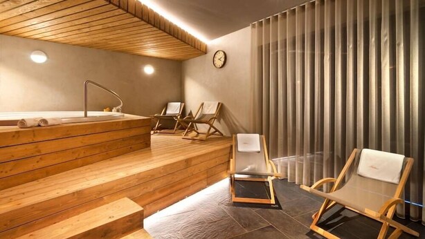 Uma área de spa moderna com escadas de madeira que levam a uma banheira de hidromassagem, duas espreguiçadeiras e uma grande janela com persianas verticais.