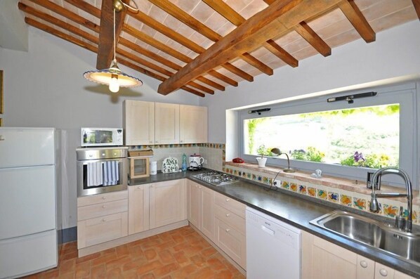 A kitchen with white cabinets, a stainless steel sink, a window with a view of greenery, and a wooden ceiling structure. - Villa Cristiana (Proceno)