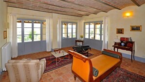 A spacious living room with a sofa, a coffee table, and a patterned rug. There are large windows with curtains, a radiator, and a wall-mounted lamp. - Villa Cristiana (Proceno)