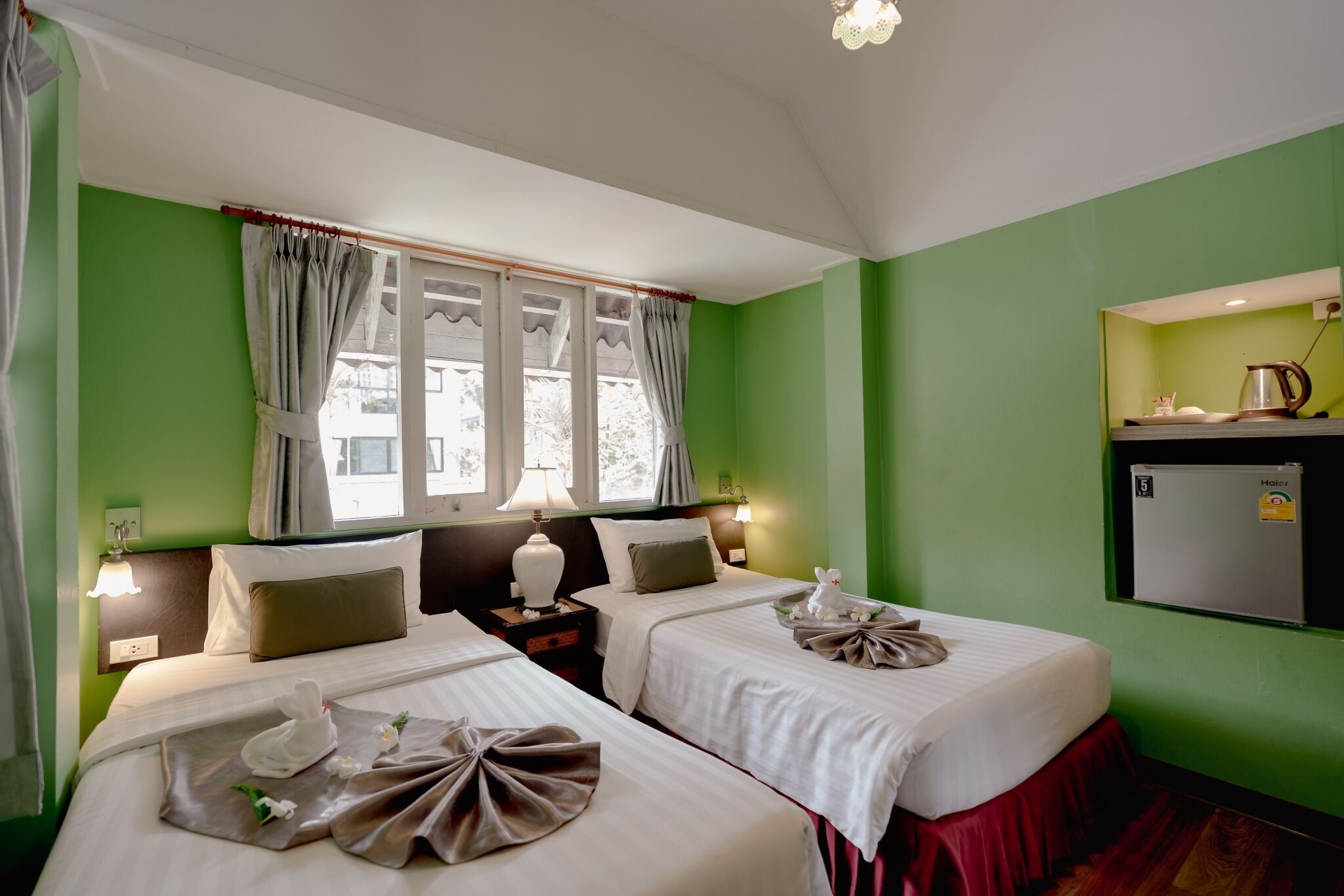 A hotel room with two beds, a window with curtains, a nightstand with a lamp, a refrigerator, and a wall-mounted shelf with a kettle.