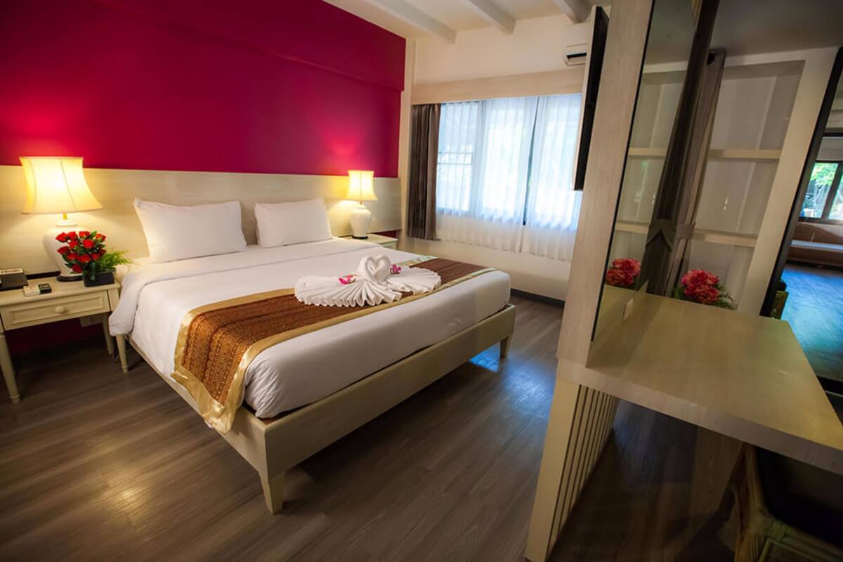 A hotel room with a large bed, two bedside tables with lamps, a red accent wall, and a view of the outdoors through a window.