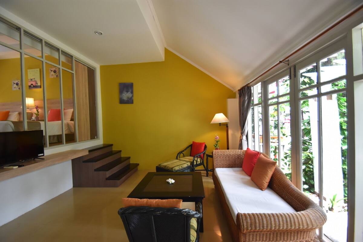 A modern living room with a sofa, armchairs, a coffee table, and a TV. There are large windows with curtains, a staircase, and a yellow wall.