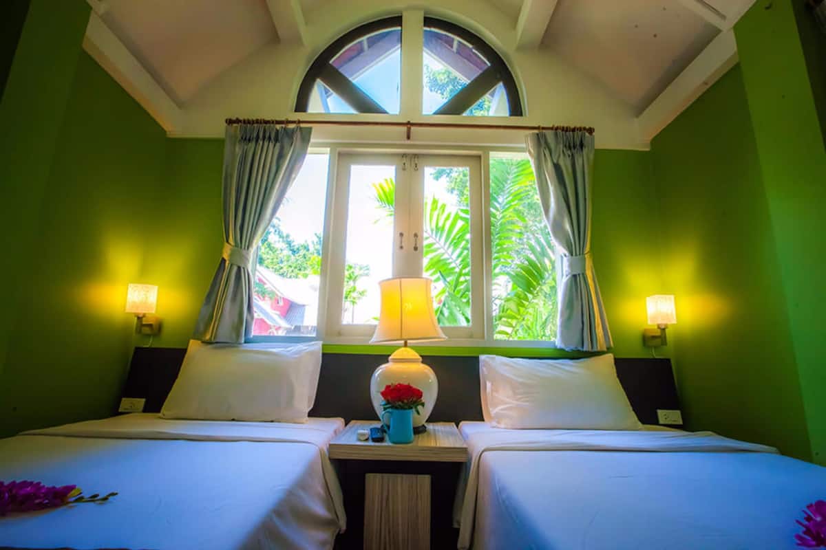 A hotel room with two beds, a window with curtains, a nightstand with a lamp, and a view of greenery outside.