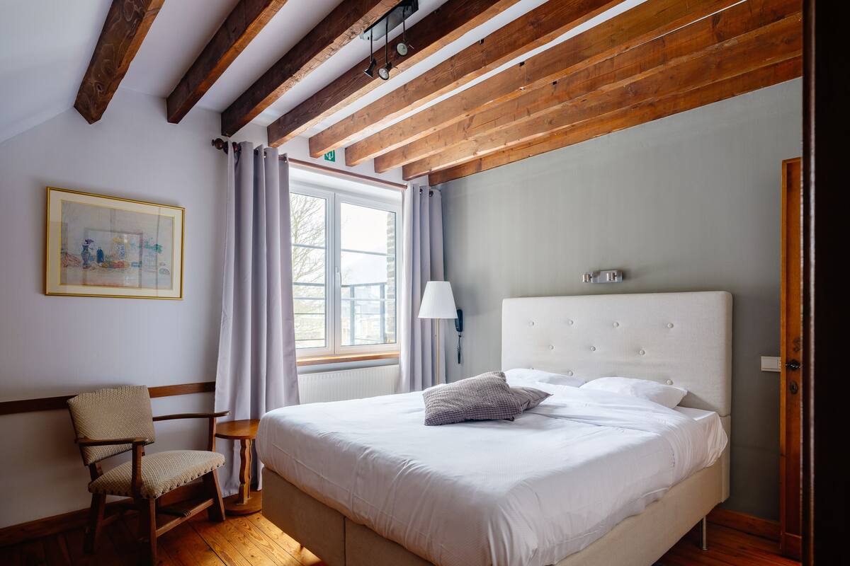 A bedroom with a large bed, wooden beams on the ceiling, a chair, a bedside table, and a window with curtains.