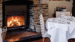 A cozy dining area with a fireplace, a table set for two, and a window with curtains. - Auberge d'Alle (Vresse-sur-Semois)
