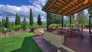 A covered patio with a table and chairs, surrounded by a well-maintained garden and tall trees. - Villa Sabrina (San Gimignano)