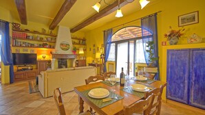A cozy living room with a fireplace, a dining area with a table set for four, and a kitchen area with blue cabinets. - Villa Sabrina (San Gimignano)