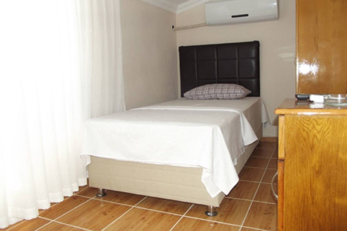 A small, neatly made bed with a headboard, a wooden nightstand, and a white curtain.
