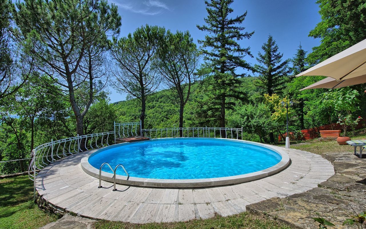 Una piscina rodona amb terrassa de fusta i barana de metall, envoltada d'arbres i un cel serè.