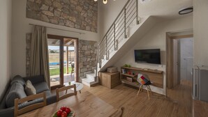 A spacious living area with a stone wall, a wooden staircase, a flat-screen TV, and a dining table with a red fruit bowl. - Esperanza Villas (Nea Propontida)