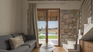 A modern living room with a grey sofa, a small white table, and a view of a pool and outdoor area through a glass door. - Esperanza Villas (Nea Propontida)