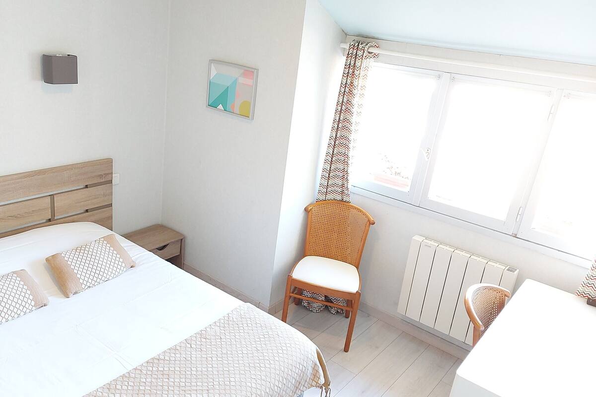 Une chambre à coucher comprenant un lit, une chaise, une table de chevet, un radiateur et une fenêtre avec des rideaux.