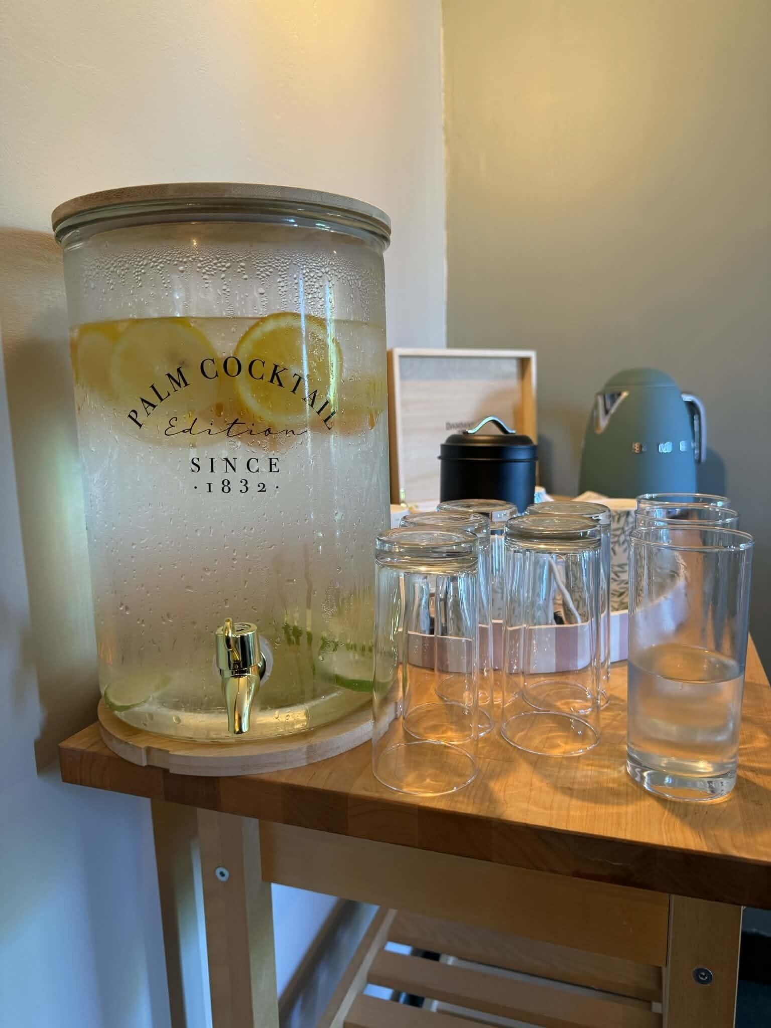 Un recipiente grande de vidrio para ponche con el rótulo 'Palm Cocktail', diseño vintage, sobre una mesa de madera.