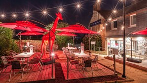 Un espace extérieur aménagé en terrasse, avec des parasols rouges, des tables et des chaises, et orné d’une sculpture de girafe originale.