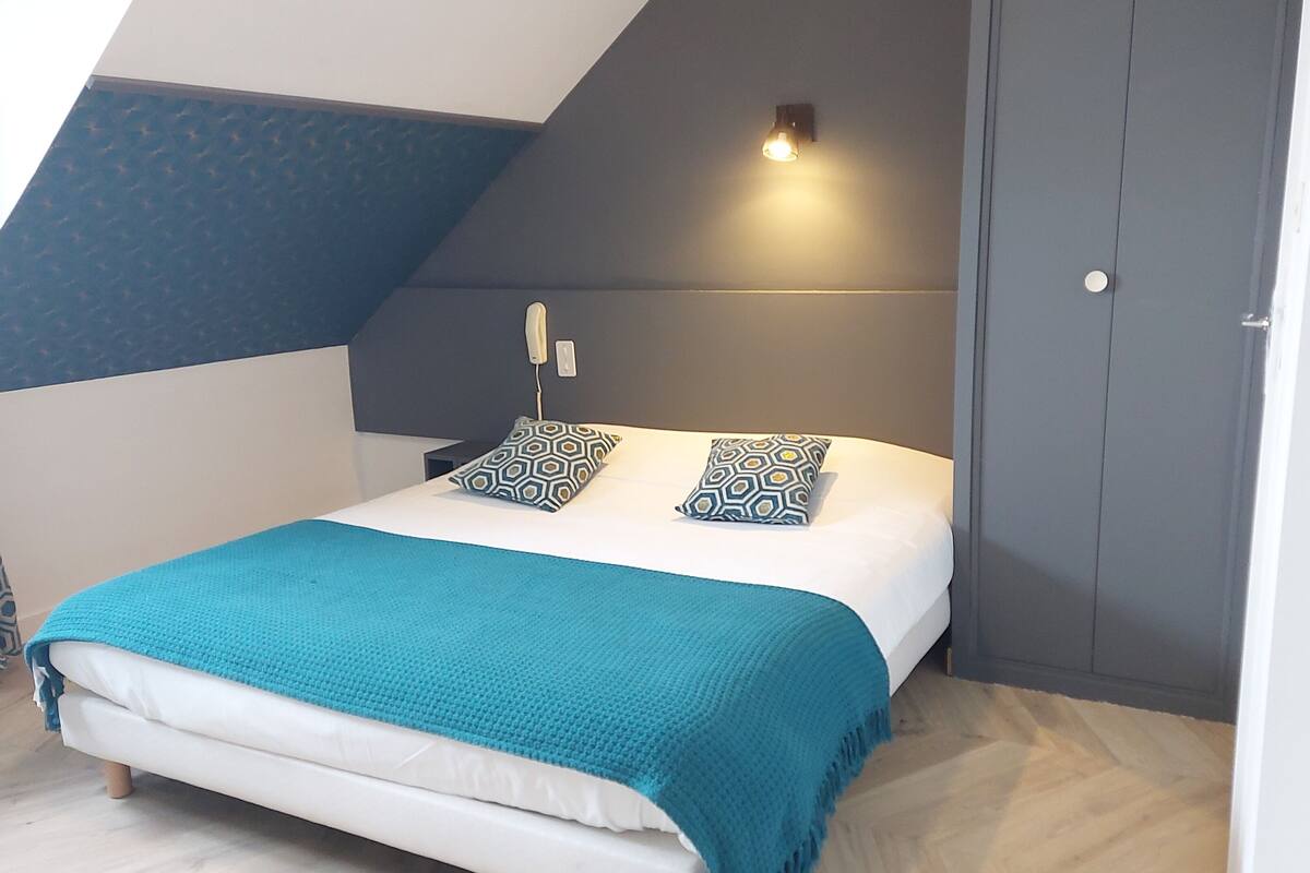 Une chambre avec un plafond en pente, un lit recouvert d’une couverture turquoise, des coussins à motifs, une lampe de chevet et une armoire.