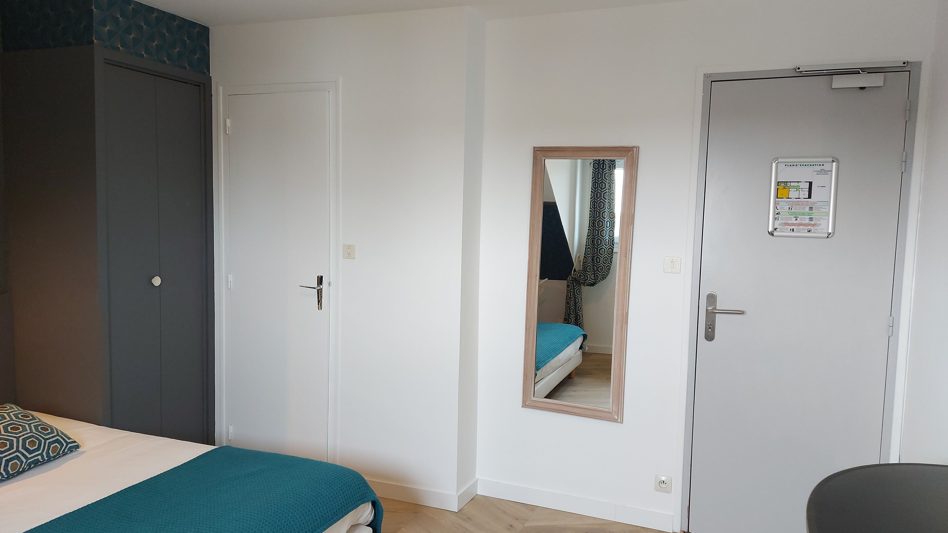 A bedroom with a bed, a wardrobe, a mirror, and a door with a fire evacuation sign.