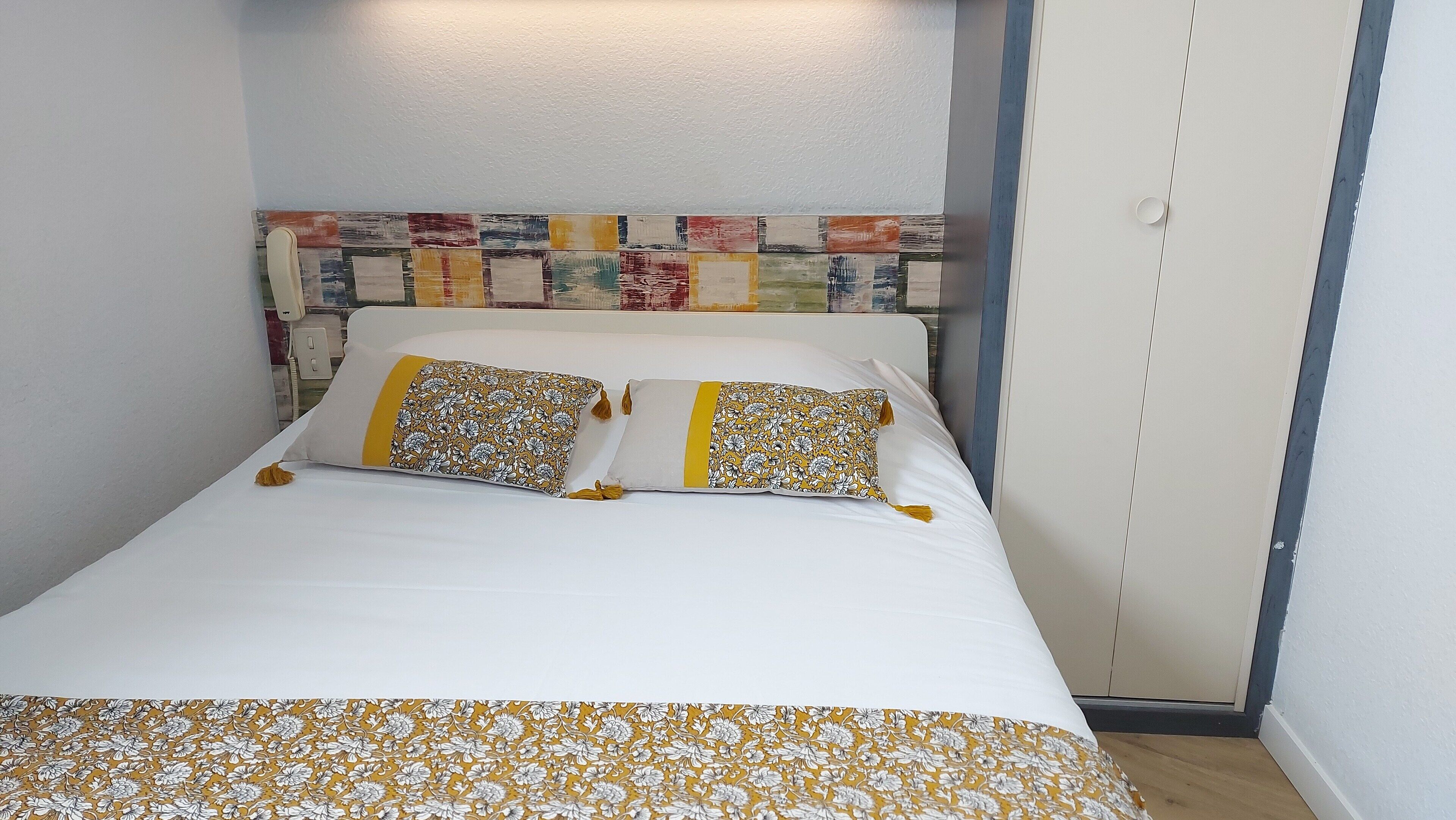 Una cama bien hecha con ropa de cama de estampado floral y dos almohadas, un cabecero de madera con un diseño colorido de patchwork y un armario blanco a la derecha.