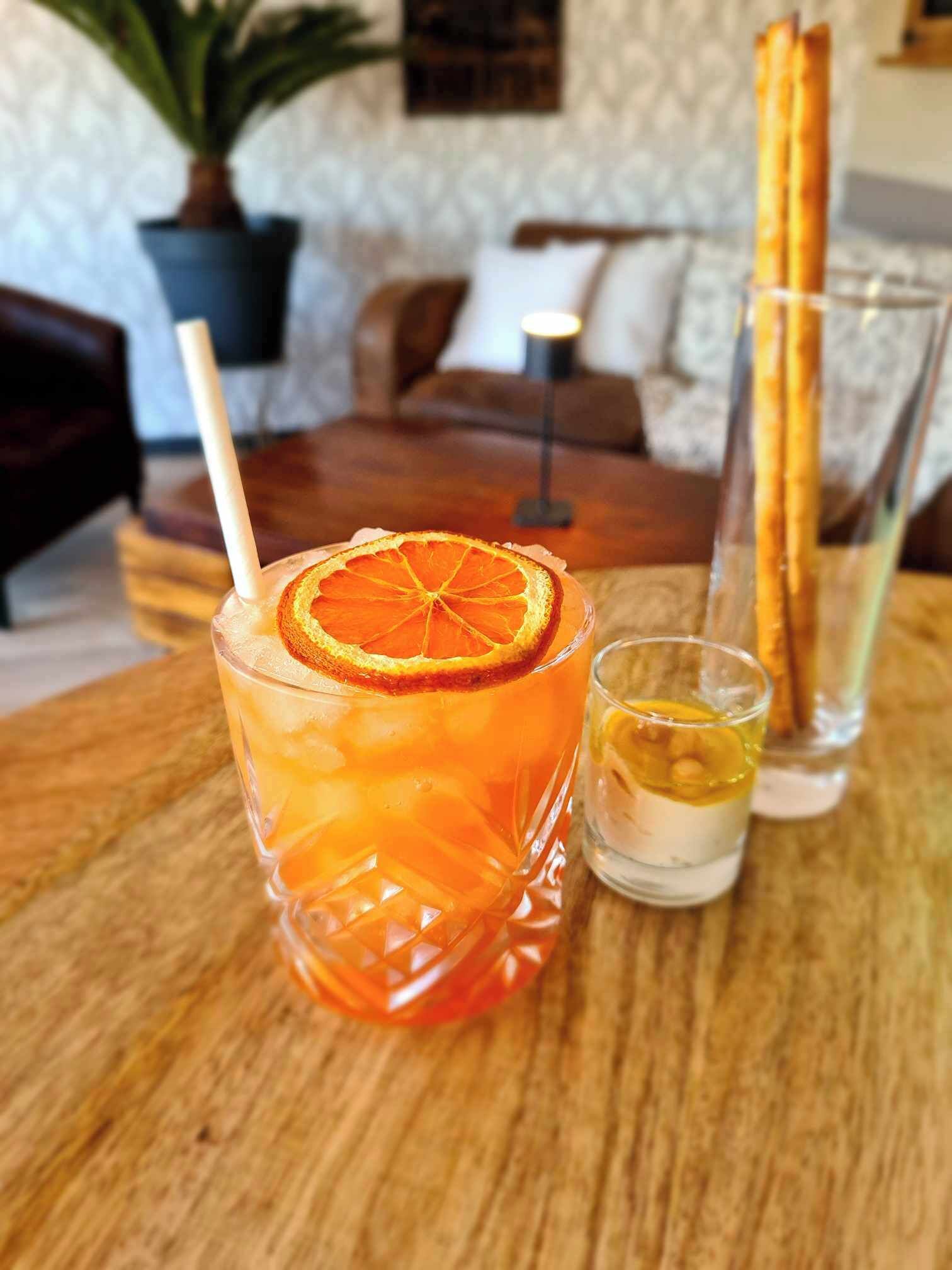 Um copo de suco de laranja com uma fatia de laranja na borda, um copo medidor com líquido amarelado e um copo de água com uma fatia de limão sobre uma mesa de madeira.
