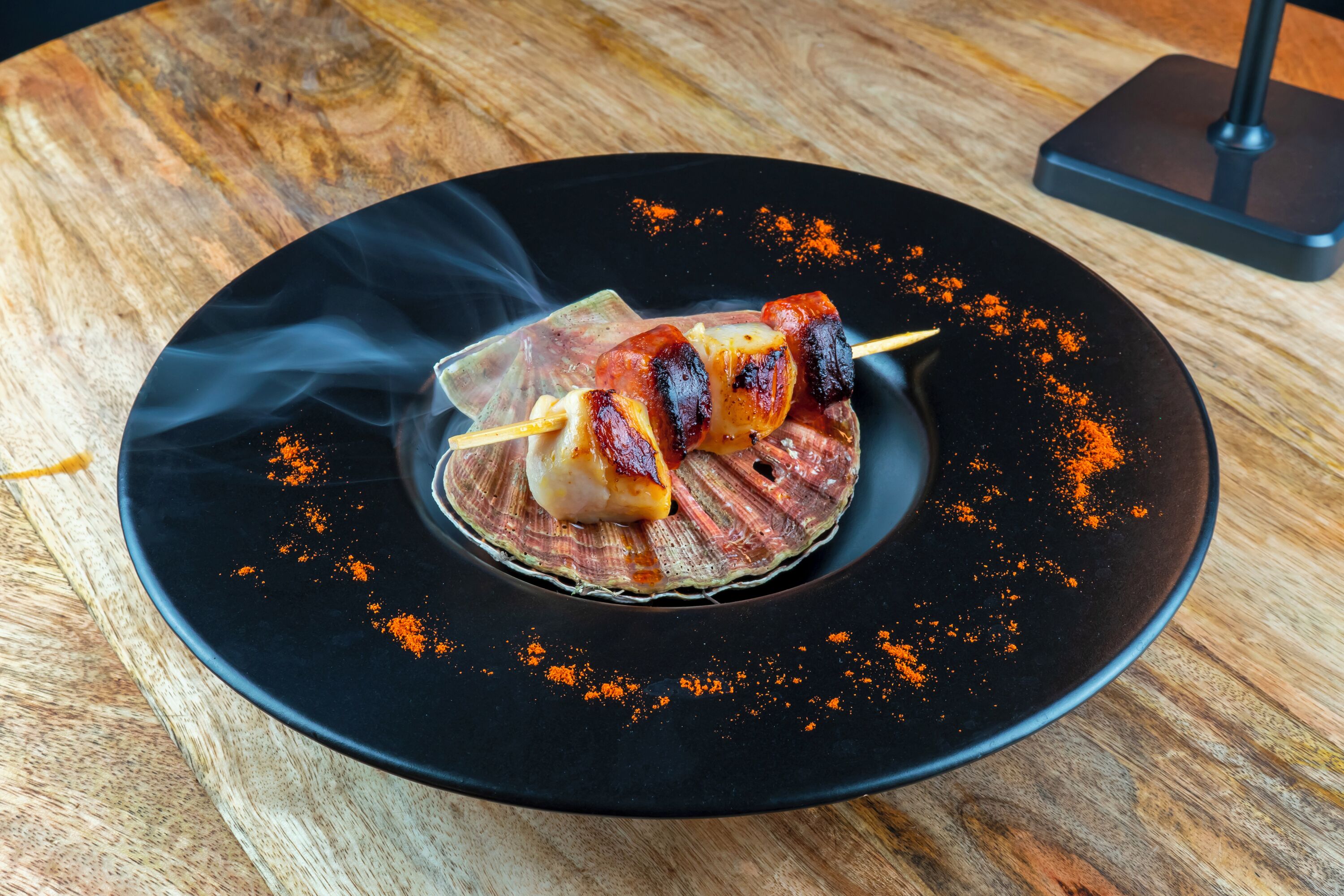 Une assiette avec des aliments présentés sur des brochettes, une assiette noire et une table en bois.