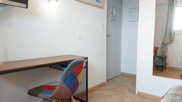 Una habitación pequeña y escasamente amoblada, con un escritorio de madera, una silla colorida, una puerta y un espejo.