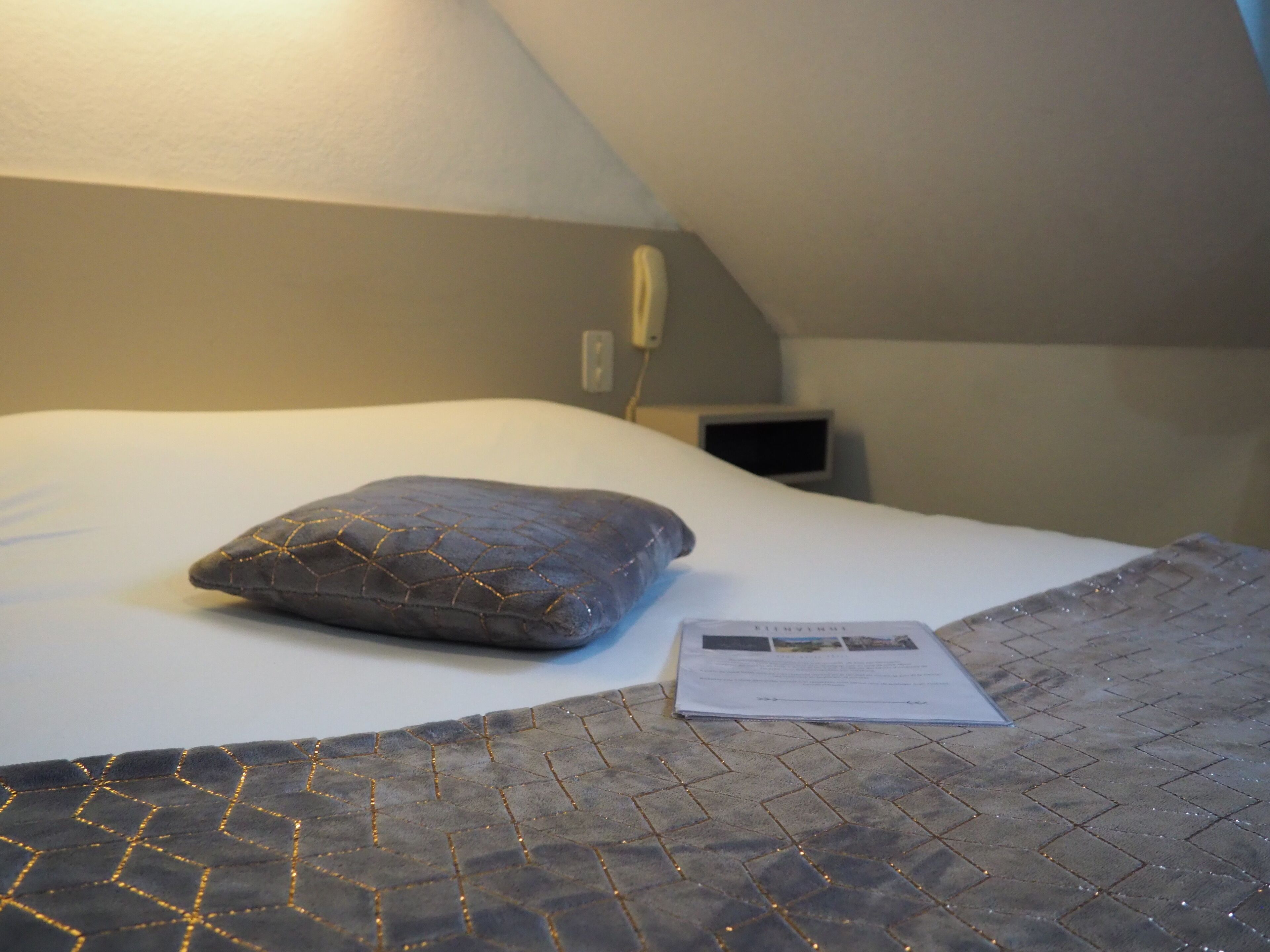 Un dormitorio con cama, almohada, mesita de noche y enchufe en la pared.